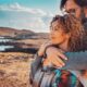 A couple embraces outdoors by the ocean at sunset, with the woman looking ahead thoughtfully while the man gently holds her from behind, symbolizing intimacy, connection, and comfort.