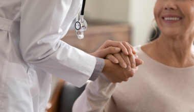 Doctor giving hope. Close up shot of young female physician leaning forward to smiling elderly lady patient holding her hand in palms. Woman caretaker in white coat supporting encouraging old person. How to talk to your doctor about perimenopause symptoms and treatment goals