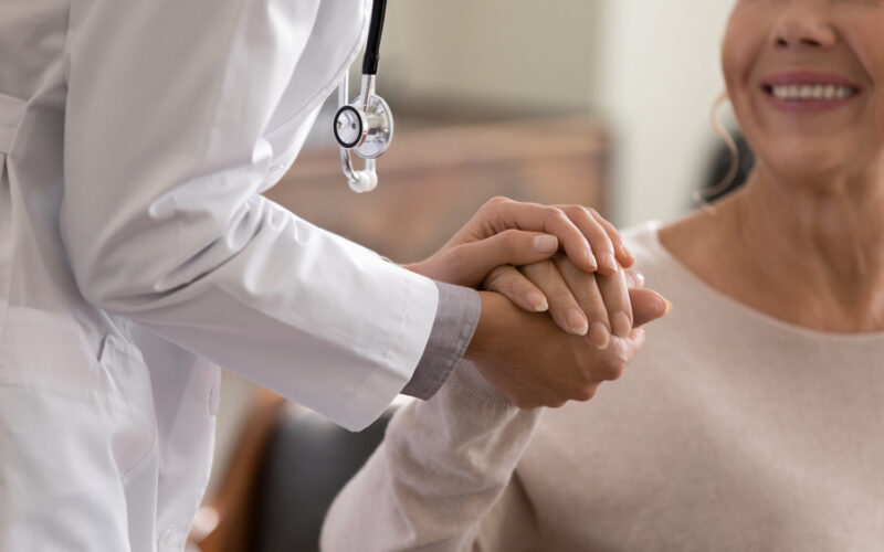 Doctor giving hope. Close up shot of young female physician leaning forward to smiling elderly lady patient holding her hand in palms. Woman caretaker in white coat supporting encouraging old person. How to talk to your doctor about perimenopause symptoms and treatment goals