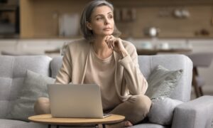 A thoughtful middle-aged woman with gray hair sits cross-legged on a sofa with a laptop in front of her, resting her chin on her hand as she looks away, appearing reflective.
