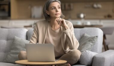 A thoughtful middle-aged woman with gray hair sits cross-legged on a sofa with a laptop in front of her, resting her chin on her hand as she looks away, appearing reflective.