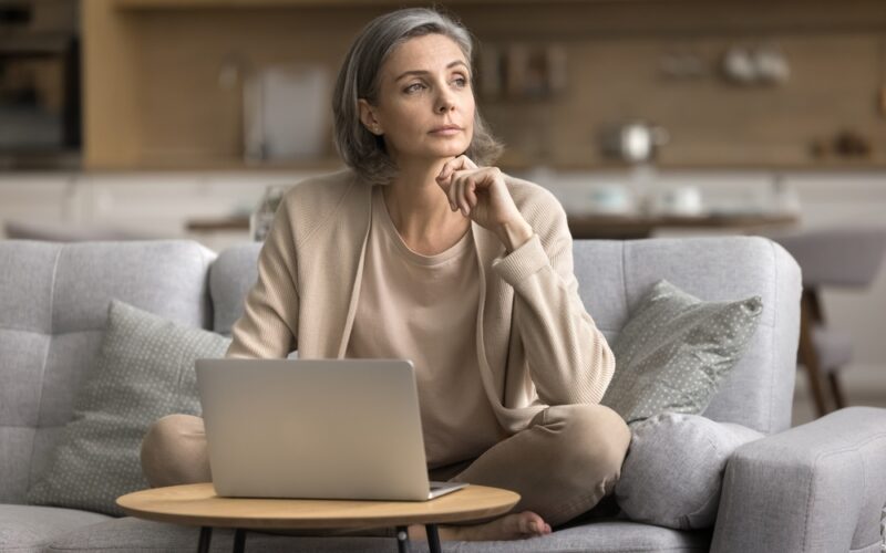 A thoughtful middle-aged woman with gray hair sits cross-legged on a sofa with a laptop in front of her, resting her chin on her hand as she looks away, appearing reflective.