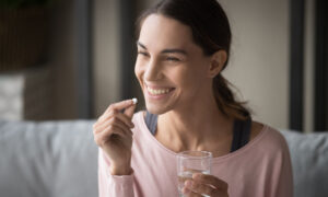 Smiling healthy young woman holding pill glass of water sit on sofa at home, positive lady take daily medicine antioxidant diet vitamin supplements for beauty skin hair health care medicament. Sermorelin vs traditional hormone replacement therapy