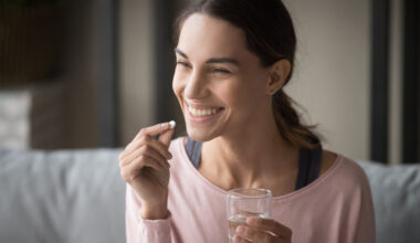 Smiling healthy young woman holding pill glass of water sit on sofa at home, positive lady take daily medicine antioxidant diet vitamin supplements for beauty skin hair health care medicament. Sermorelin vs traditional hormone replacement therapy