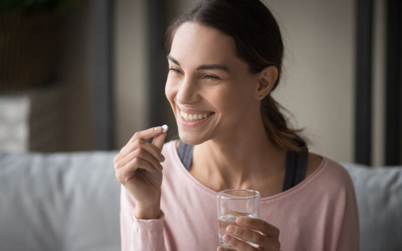 Smiling healthy young woman holding pill glass of water sit on sofa at home, positive lady take daily medicine antioxidant diet vitamin supplements for beauty skin hair health care medicament. Sermorelin vs traditional hormone replacement therapy
