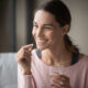 Smiling healthy young woman holding pill glass of water sit on sofa at home, positive lady take daily medicine antioxidant diet vitamin supplements for beauty skin hair health care medicament. Sermorelin vs traditional hormone replacement therapy