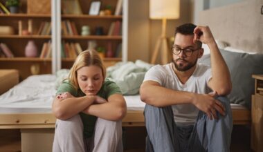 A couple sits on the floor by their bed, both looking down and appearing withdrawn, symbolizing the emotional impact of mental health challenges on intimacy and sexual relationships.