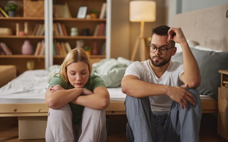 A couple sits on the floor by their bed, both looking down and appearing withdrawn, symbolizing the emotional impact of mental health challenges on intimacy and sexual relationships.