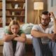 A couple sits on the floor by their bed, both looking down and appearing withdrawn, symbolizing the emotional impact of mental health challenges on intimacy and sexual relationships.