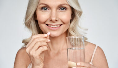Smiling happy healthy middle aged 50s woman holding glass of water taking dietary supplement vitamin pink pill isolated on white background. Old women multivitamins antioxidants for anti age beauty.