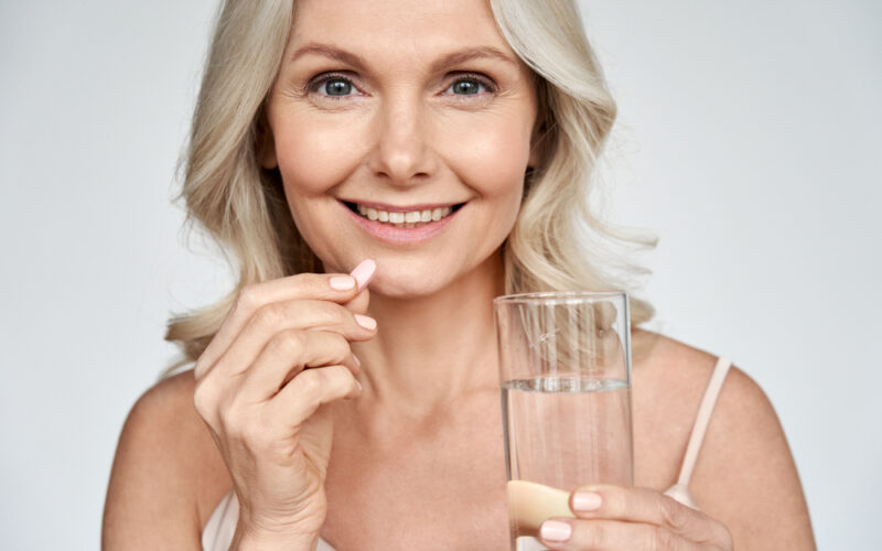 Smiling happy healthy middle aged 50s woman holding glass of water taking dietary supplement vitamin pink pill isolated on white background. Old women multivitamins antioxidants for anti age beauty.