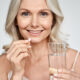 Smiling happy healthy middle aged 50s woman holding glass of water taking dietary supplement vitamin pink pill isolated on white background. Old women multivitamins antioxidants for anti age beauty.