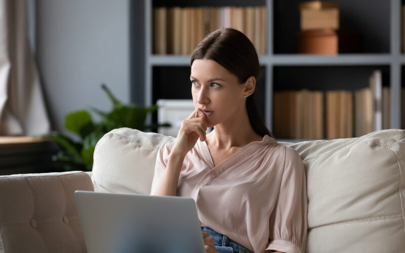 Woman sitting on a couch with a laptop, thoughtfully considering acarbose costs and whether the medication is worth it.