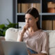 Woman sitting on a couch with a laptop, thoughtfully considering acarbose costs and whether the medication is worth it.