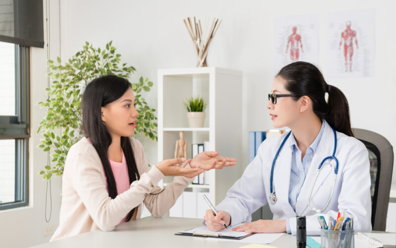 A woman talks openly with her female doctor during a medical consultation in a bright office. The doctor, wearing a white coat and stethoscope, listens attentively and takes notes, symbolizing supportive care for sensitive health concerns.