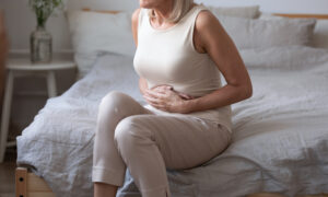 Older woman taking tirzepatide sitting on bed holding her stomach, appearing uncomfortable from abdominal pain or diarrhea.