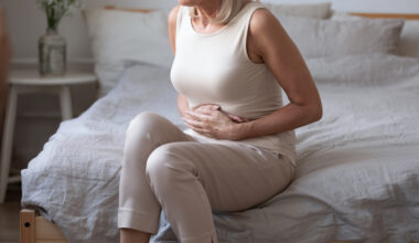 Older woman taking tirzepatide sitting on bed holding her stomach, appearing uncomfortable from abdominal pain or diarrhea.