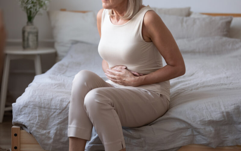 Older woman taking tirzepatide sitting on bed holding her stomach, appearing uncomfortable from abdominal pain or diarrhea.