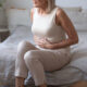 Older woman taking tirzepatide sitting on bed holding her stomach, appearing uncomfortable from abdominal pain or diarrhea.