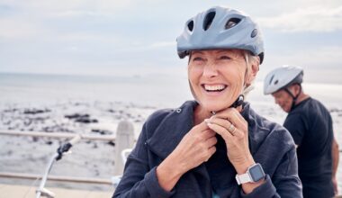 A smiling older woman fastens her bike helmet while standing near the ocean, with another cyclist in the background, symbolizing energy, vitality, and healthy aging.