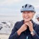 A smiling older woman fastens her bike helmet while standing near the ocean, with another cyclist in the background, symbolizing energy, vitality, and healthy aging.