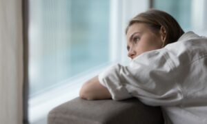 A woman leans on the back of a couch, gazing thoughtfully out a large window with a pensive expression, symbolizing the emotional impact of persistent low sexual desire.