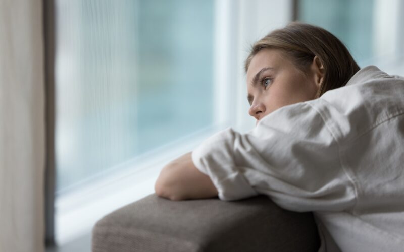 A woman leans on the back of a couch, gazing thoughtfully out a large window with a pensive expression, symbolizing the emotional impact of persistent low sexual desire.