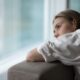A woman leans on the back of a couch, gazing thoughtfully out a large window with a pensive expression, symbolizing the emotional impact of persistent low sexual desire.