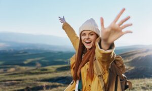 A joyful young woman in a yellow jacket and beanie raises her arms in celebration while hiking in the mountains, symbolizing energy, vitality, and wellness.
