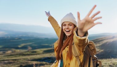 A joyful young woman in a yellow jacket and beanie raises her arms in celebration while hiking in the mountains, symbolizing energy, vitality, and wellness.