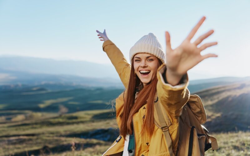 A joyful young woman in a yellow jacket and beanie raises her arms in celebration while hiking in the mountains, symbolizing energy, vitality, and wellness.