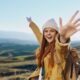 A joyful young woman in a yellow jacket and beanie raises her arms in celebration while hiking in the mountains, symbolizing energy, vitality, and wellness.