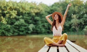 Young woman in a hat and yellow pants sitting cross-legged on a wooden dock by the water, smiling and raising her arms joyfully, symbolizing energy, balance, and vitality associated with NAD⁺ IV therapy.