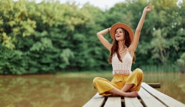 Young woman in a hat and yellow pants sitting cross-legged on a wooden dock by the water, smiling and raising her arms joyfully, symbolizing energy, balance, and vitality associated with NAD⁺ IV therapy.