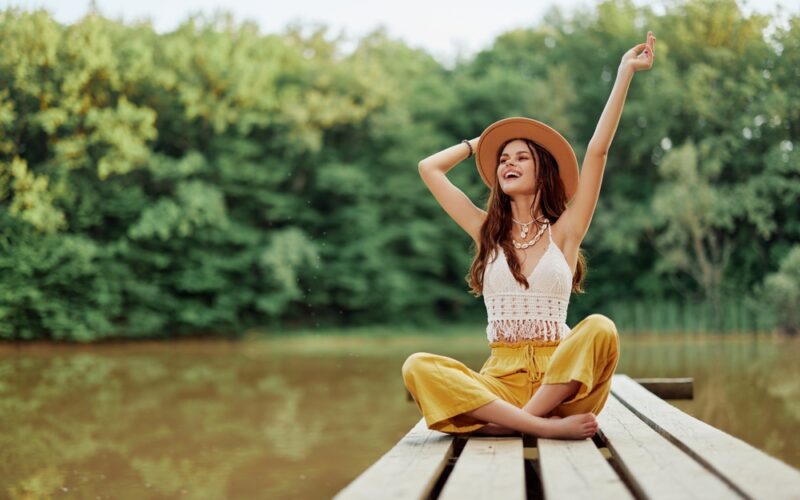 Young woman in a hat and yellow pants sitting cross-legged on a wooden dock by the water, smiling and raising her arms joyfully, symbolizing energy, balance, and vitality associated with NAD⁺ IV therapy.