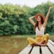 Young woman in a hat and yellow pants sitting cross-legged on a wooden dock by the water, smiling and raising her arms joyfully, symbolizing energy, balance, and vitality associated with NAD⁺ IV therapy.