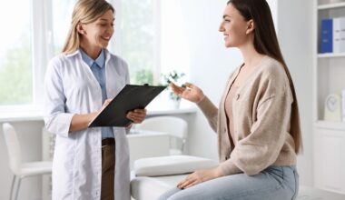 A smiling young woman sits on an exam table speaking with a female doctor who holds a clipboard, symbolizing open communication about reproductive and vaginal health concerns.