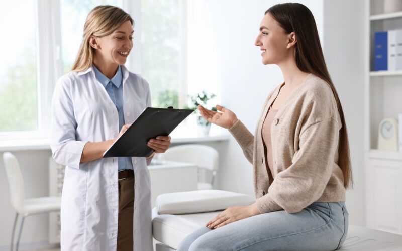 A smiling young woman sits on an exam table speaking with a female doctor who holds a clipboard, symbolizing open communication about reproductive and vaginal health concerns.