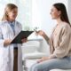 A smiling young woman sits on an exam table speaking with a female doctor who holds a clipboard, symbolizing open communication about reproductive and vaginal health concerns.