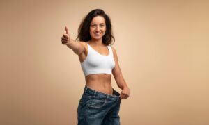Smiling woman in a white sports bra giving a thumbs-up while holding out the waistband of her loose jeans, symbolizing successful weight loss with Zepbound (tirzepatide).