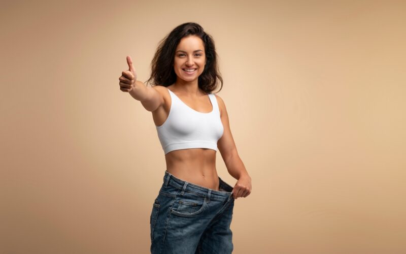 Smiling woman in a white sports bra giving a thumbs-up while holding out the waistband of her loose jeans, symbolizing successful weight loss with Zepbound (tirzepatide).