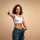 Smiling woman in a white sports bra giving a thumbs-up while holding out the waistband of her loose jeans, symbolizing successful weight loss with Zepbound (tirzepatide).