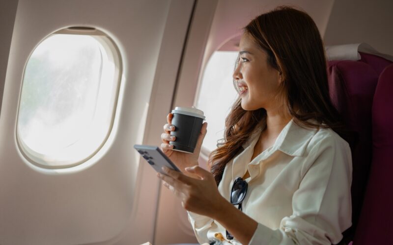 Woman traveling with tirzepatide, holding a coffee cup and phone while sitting on an airplane.