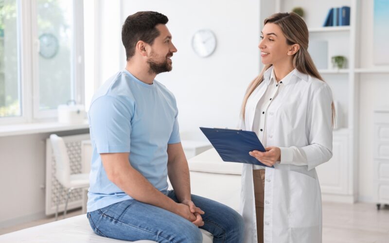 Male patient talking with a female doctor holding a clipboard during a consultation, representing medical guidance on vitamin B12 injections and their role in weight management.