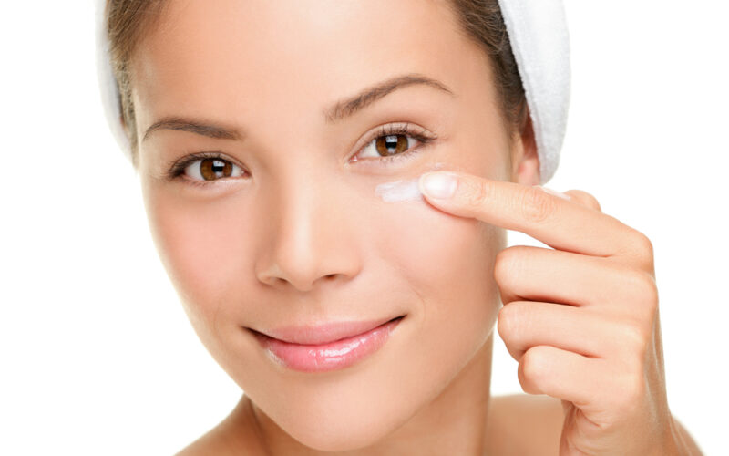 Close-up of a smiling woman applying cream under her eye with her fingertip, representing skincare and the cautious use of tretinoin in the delicate under-eye area.