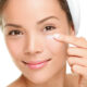 Close-up of a smiling woman applying cream under her eye with her fingertip, representing skincare and the cautious use of tretinoin in the delicate under-eye area.