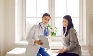 Female doctor sitting beside a smiling woman in a bright medical office, reviewing health information on a clipboard — representing patient guidance on managing acarbose side effects and improving comfort during diabetes treatment.