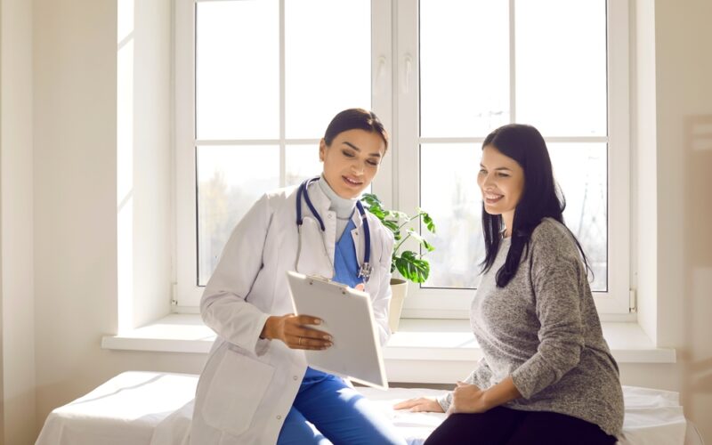 Female doctor sitting beside a smiling woman in a bright medical office, reviewing health information on a clipboard — representing patient guidance on managing acarbose side effects and improving comfort during diabetes treatment.
