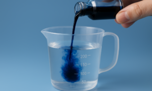 Close-up of a dark blue liquid being poured from a small glass bottle into a measuring cup containing 200 mL of clear water, illustrating how to properly dilute methylene blue for safe oral use.