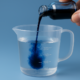 Close-up of a dark blue liquid being poured from a small glass bottle into a measuring cup containing 200 mL of clear water, illustrating how to properly dilute methylene blue for safe oral use.